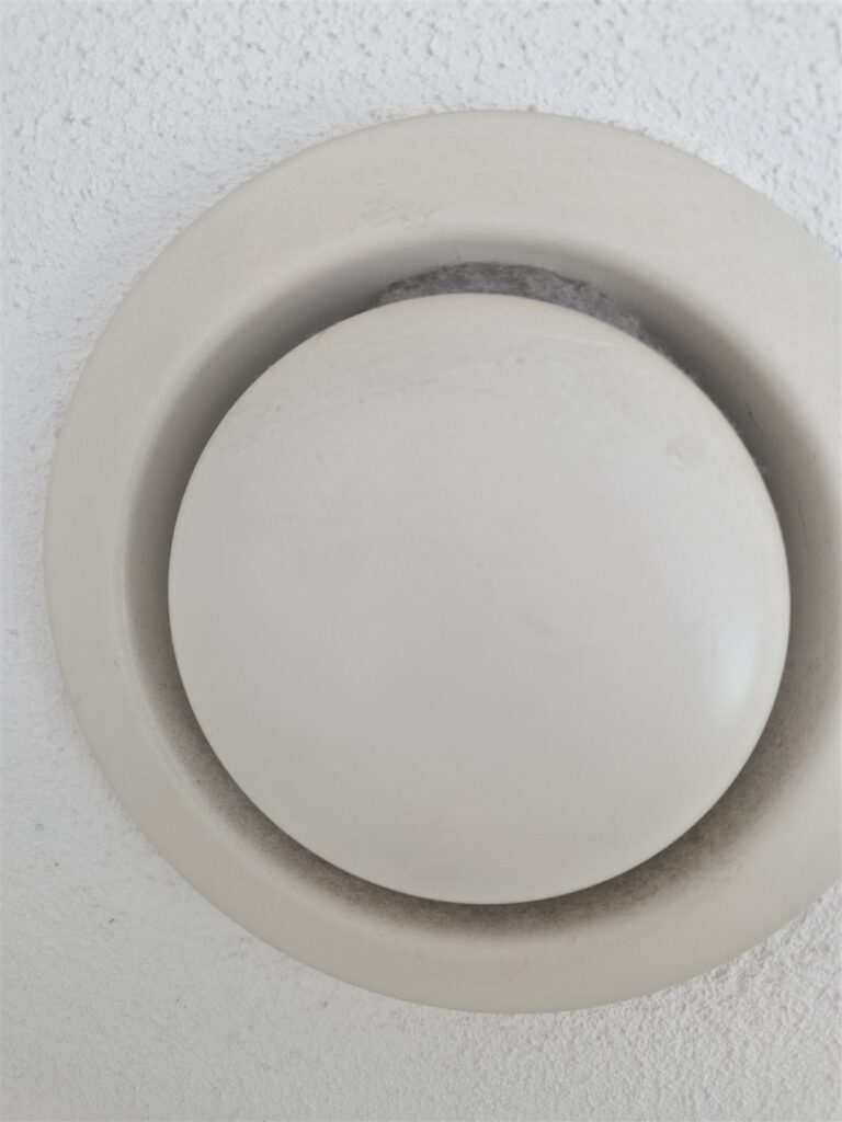 Weißes Tellerventil einer Wohnraumlüftung mit sichtbaren Staubablagerungen am Luftspalt.